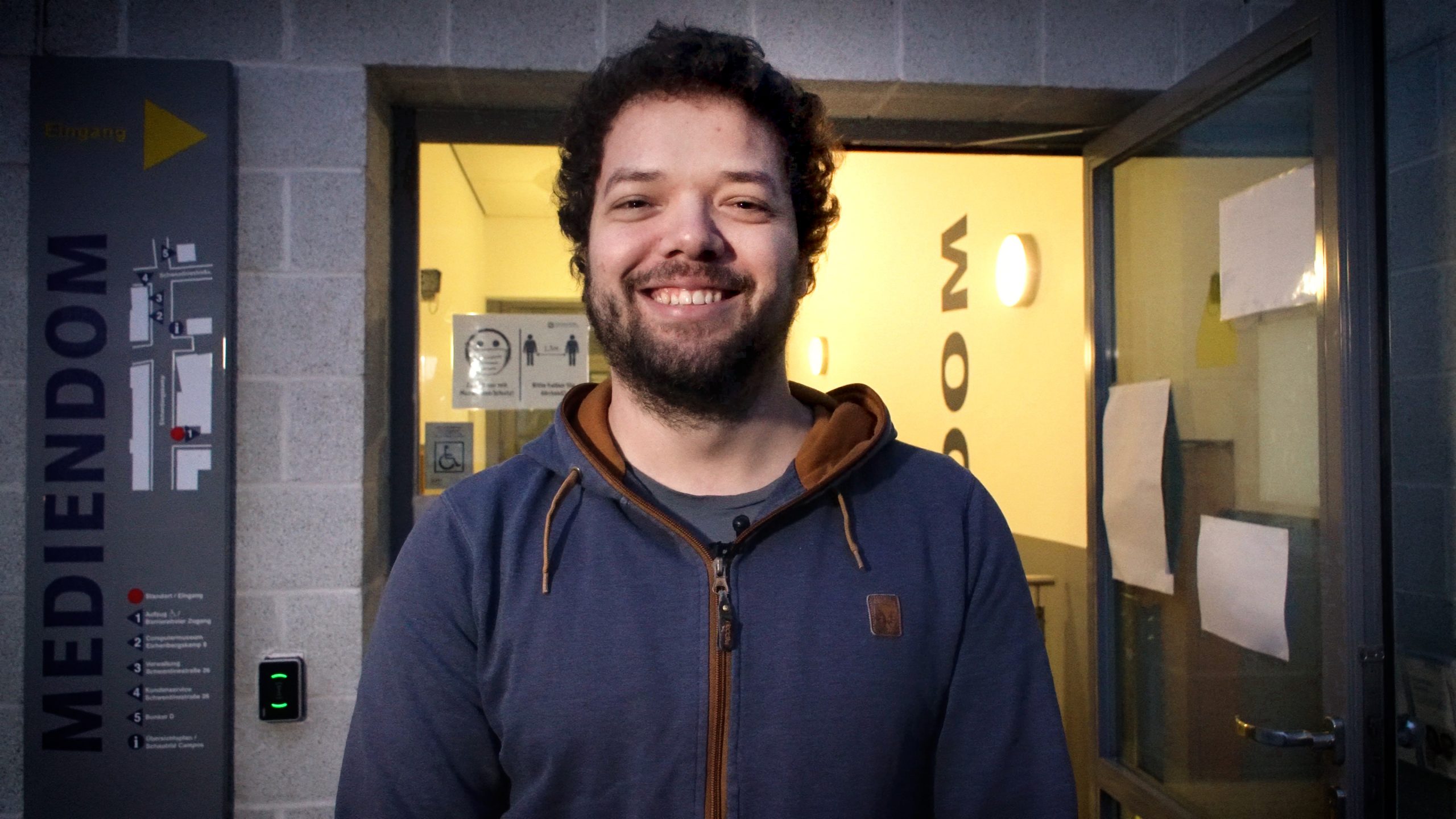 Lächelnder Mann mit dunklem Bart und Haar vor dem Eingang des Mediendoms.