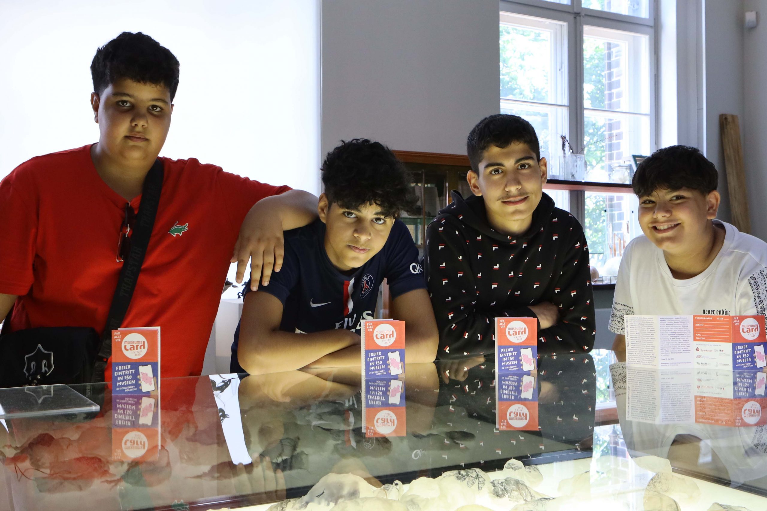 Vier Teenager in einem Museum über einem Ausstellungstisch mit Glasplatte und vor ihnen der Flyer der Museumscard.